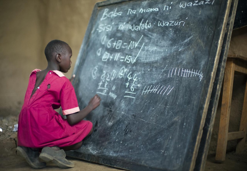 A girl in a red dress working on arithmetic with chalk at a classroom blackboard.
